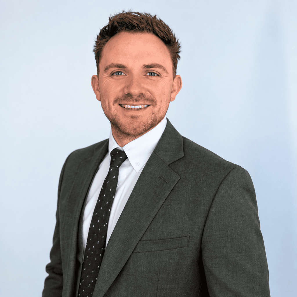 Professional man in a grey suit smiling against a light blue background.